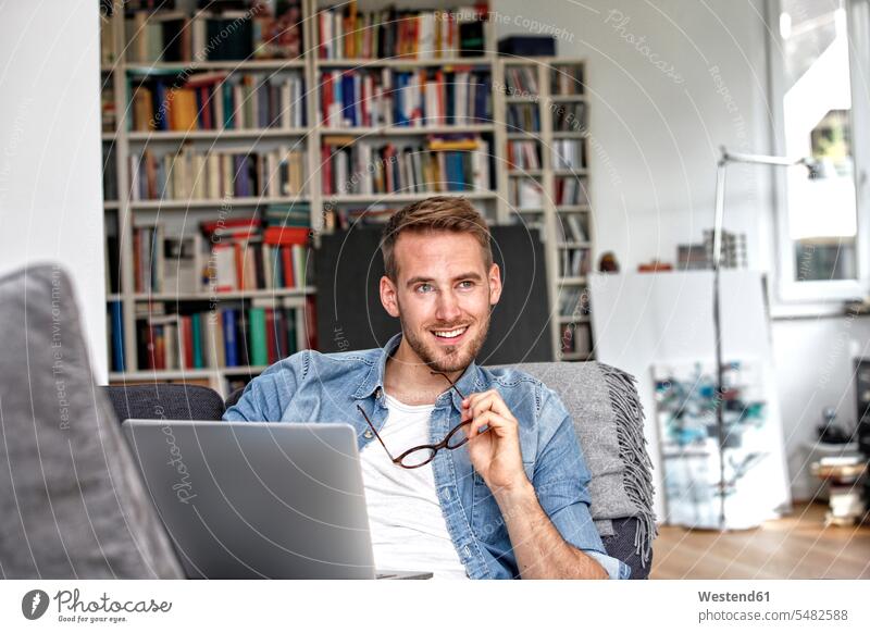 Porträt eines lächelnden Mannes, der auf einer Couch mit Laptop im Wohnzimmer sitzt Notebook Laptops Notebooks Männer männlich Computer Rechner Erwachsener