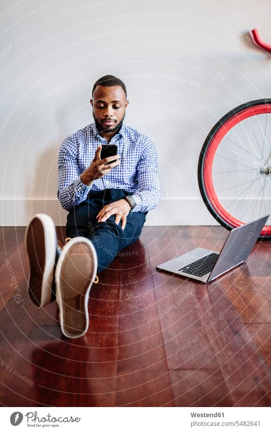 Mann auf Holzboden sitzend mit Laptop und Handy und Fahrrad neben sich Geschäftsmann Businessmann Businessmänner Geschäftsmänner sitzt Mobiltelefon Handies