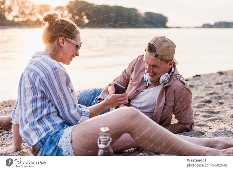 Junges Paar entspannt sich am Flussufer entspanntheit relaxt Handy Mobiltelefon Handies Handys Mobiltelefone lächeln Pärchen Paare Partnerschaft Entspannung