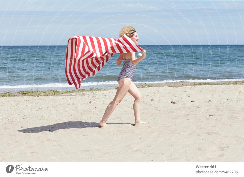 Lächelnde junge Frau mit Strandhandtuch vor dem Meer weiblich Frauen Strandtuch Beach Straende Strände Beaches Erwachsener erwachsen Mensch Menschen Leute