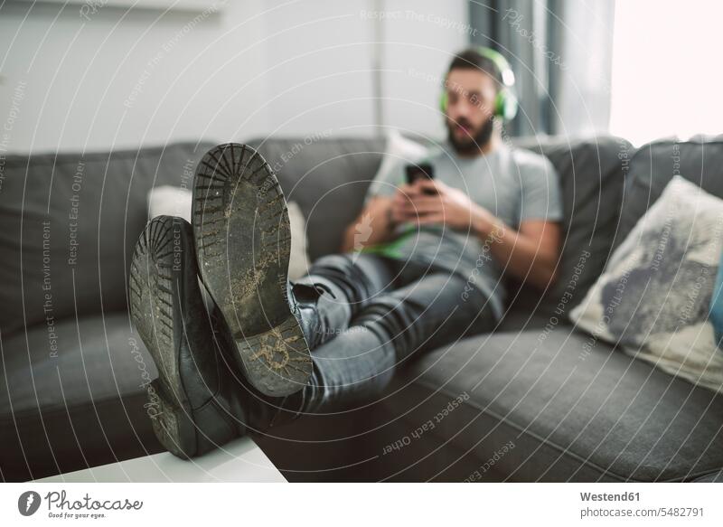 Junger Mann sitzt auf dem Sofa und hört Musik von seinem Smartphone jung Männer männlich Musik hören Kopfhörer Kopfhoerer Handy Mobiltelefon Handies Handys