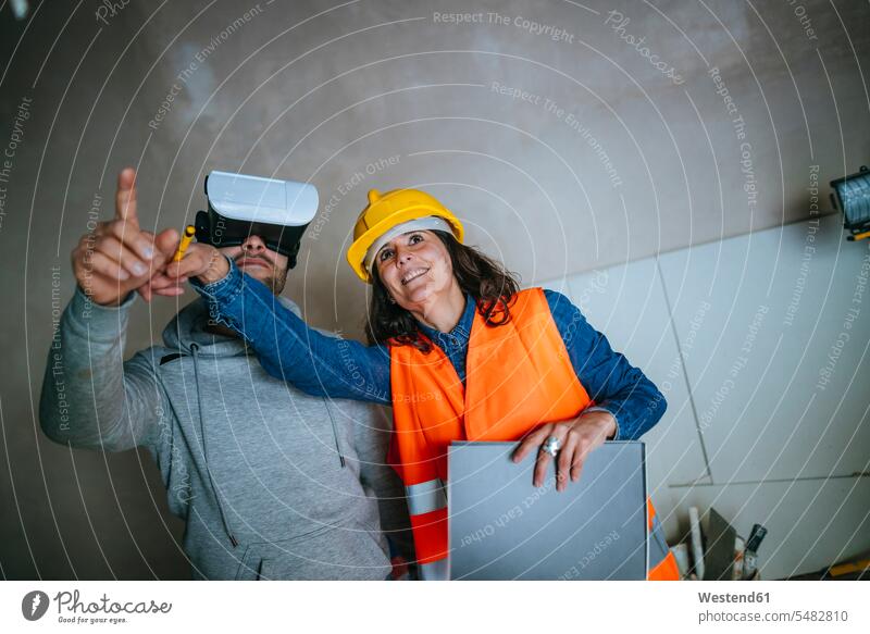 Frau auf der Baustelle zeigt einem Mann mit einer Virtual-Reality-Brille das Haus Notizblock Notizbloecke Notizblöcke arbeiten Arbeit Baustellen zeigen