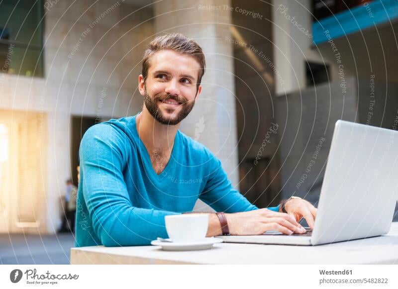 Lächelnder junger Mann sitzt mit Laptop am Tisch Notebook Laptops Notebooks Männer männlich lächeln Computer Rechner Erwachsener erwachsen Mensch Menschen Leute