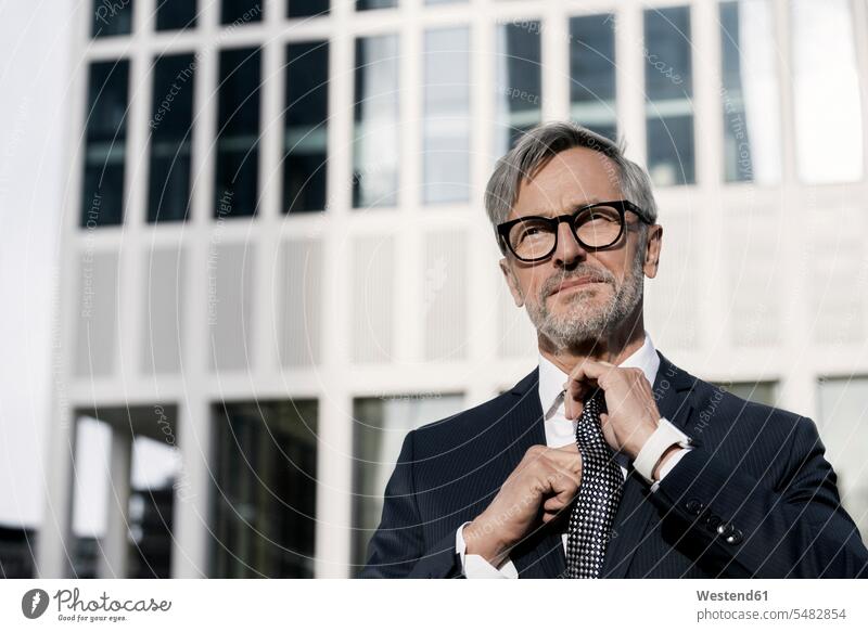 Grauhaariger Geschäftsmann richtet seine Krawatte vor einem Wolkenkratzer Businessmann Businessmänner Geschäftsmänner Krawatten Schlipse Portrait Porträts
