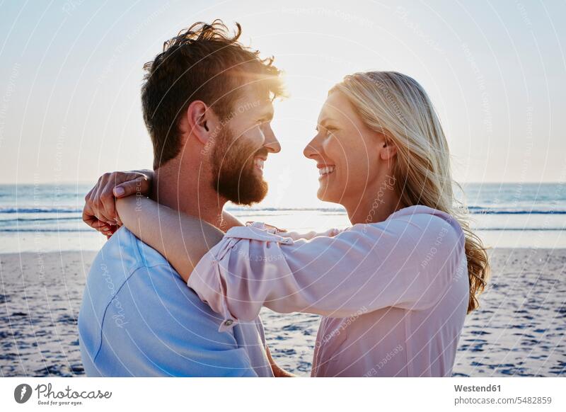 Lächelndes Paar umarmt sich am Strand bei Sonnenuntergang lächeln umarmen Umarmung Umarmungen Arm umlegen glücklich Glück glücklich sein glücklichsein Beach