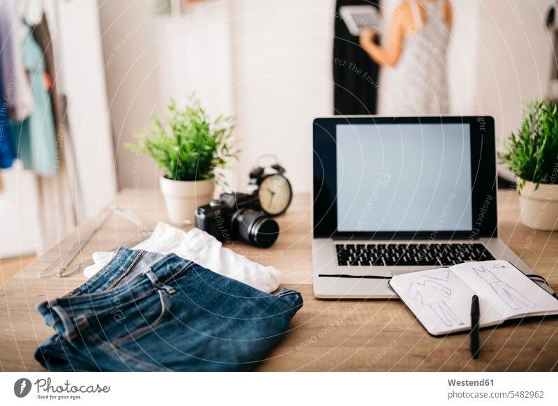 Laptop, Kleidung und Skizzenbuch auf dem Schreibtisch mit der Frau im Hintergrund Arbeitstisch Schreibtische Mode modisch Fashion Notebook Laptops Notebooks