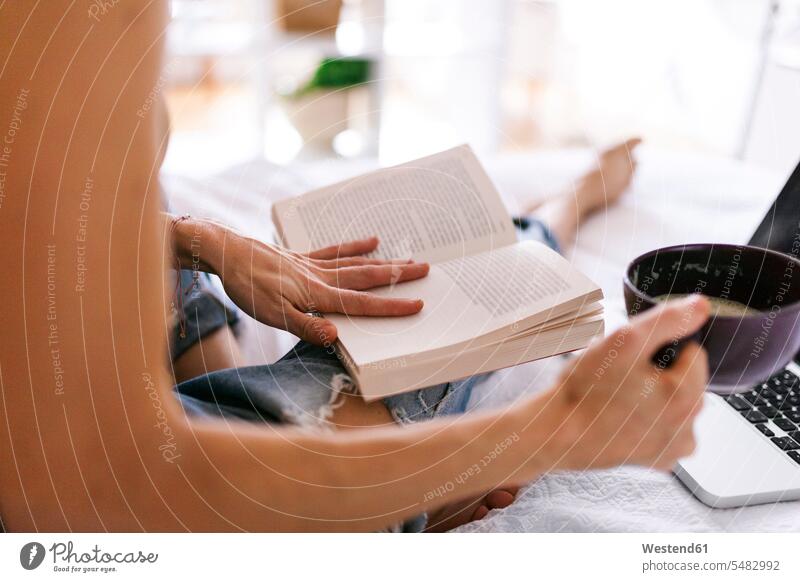 Frau sitzt im Bett, trinkt Kaffee, benutzt Laptop und liest Buch, Nahaufnahme Bücher Betten Notebook Laptops Notebooks trinken lesen Lektüre weiblich Frauen