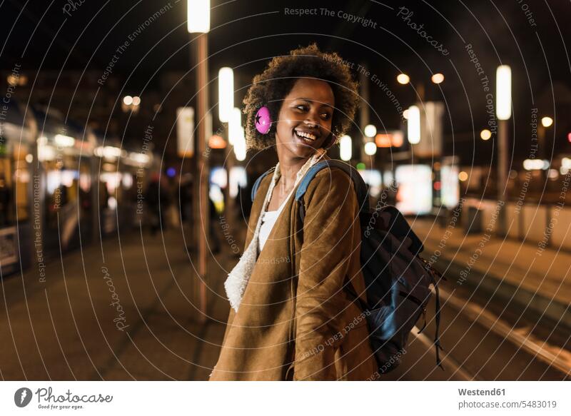 Lächelnde junge Frau mit Kopfhörern und Rucksack wartet an der Straßenbahnhaltestelle Portrait Porträts Portraits weiblich Frauen Erwachsener erwachsen Mensch