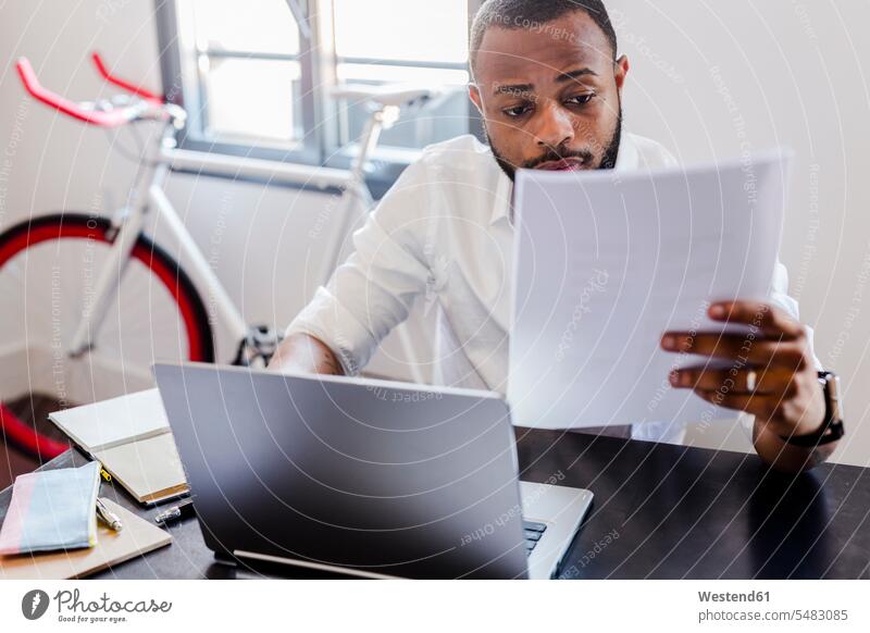 Mann betrachtet Papiere am Schreibtisch im Heimbüro Dokument Dokumente Unterlagen Männer männlich Laptop Notebook Laptops Notebooks Geschäftsmann Businessmann