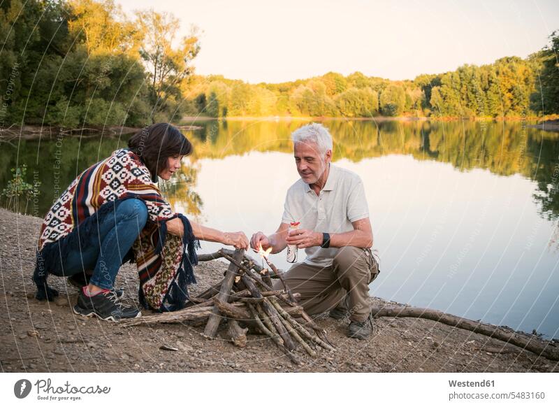 Älteres Ehepaar zündet abends ein Lagerfeuer an einem See an Senior ältere Männer älterer Mann Senioren entspannt entspanntheit relaxt Seen Paar Pärchen Paare