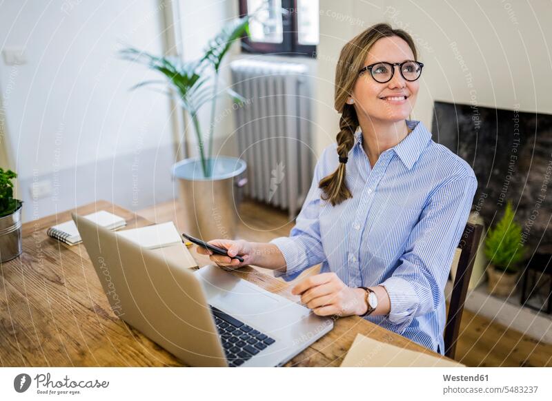 Lächelnde Frau sitzt zu Hause am Holzschreibtisch und schaut zur Seite wegsehen Blick zur Seite wegschauen Schreibtisch Arbeitstisch Schreibtische Zuhause