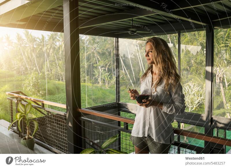 Lächelnde Frau isst Müsli in modernem Design-Haus mit Pool und Glasfassade, umgeben von einem üppigen tropischen Garten Häuser Haeuser lächeln stehen stehend