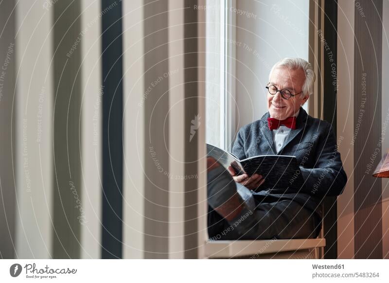 Älterer Mann sitzt auf der Fensterbank und liest Zeitschrift Senior ältere Männer älterer Mann Senioren sitzen sitzend Fensterbrett Fenstersims Fensterbänke