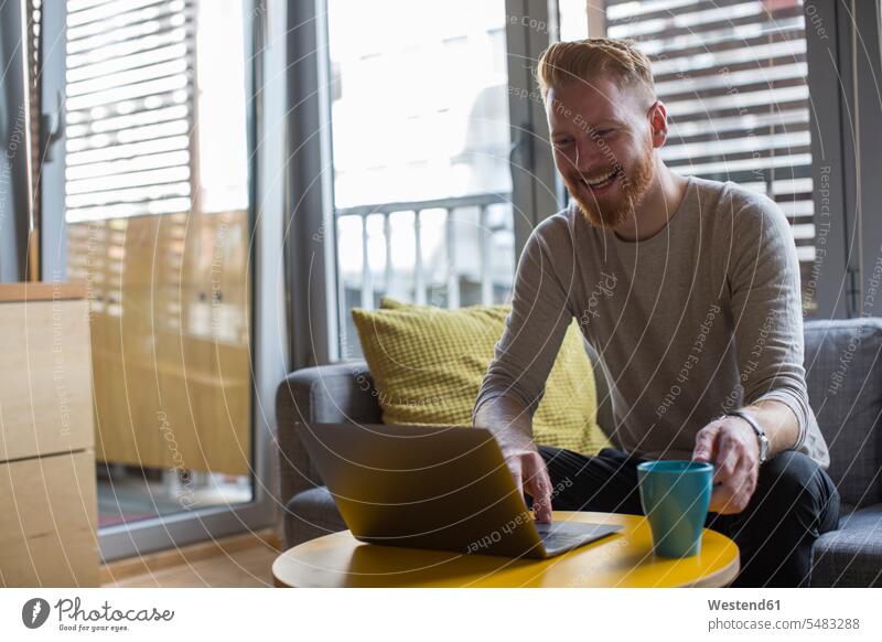 Lachender Mann mit Kaffeebecher und Laptop in seinem Wohnzimmer Notebook Laptops Notebooks Männer männlich Becher lachen Wohnraum Wohnung Wohnen Wohnräume