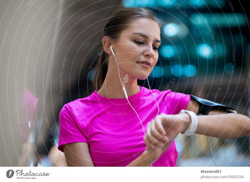 Junge Frau im rosa Sporthemd, die Musik hört und ihre Smartwatch kontrolliert Ohrhörer junge Frau junge Frauen Musik hören Joggerin Joggerinnen checken aktiv