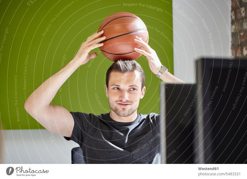 Junger Mann arbeitet im Büro und balanciert einen Basketball Portrait Porträts Portraits arbeiten Arbeit sitzen sitzend sitzt Ball Bälle balancieren Balance