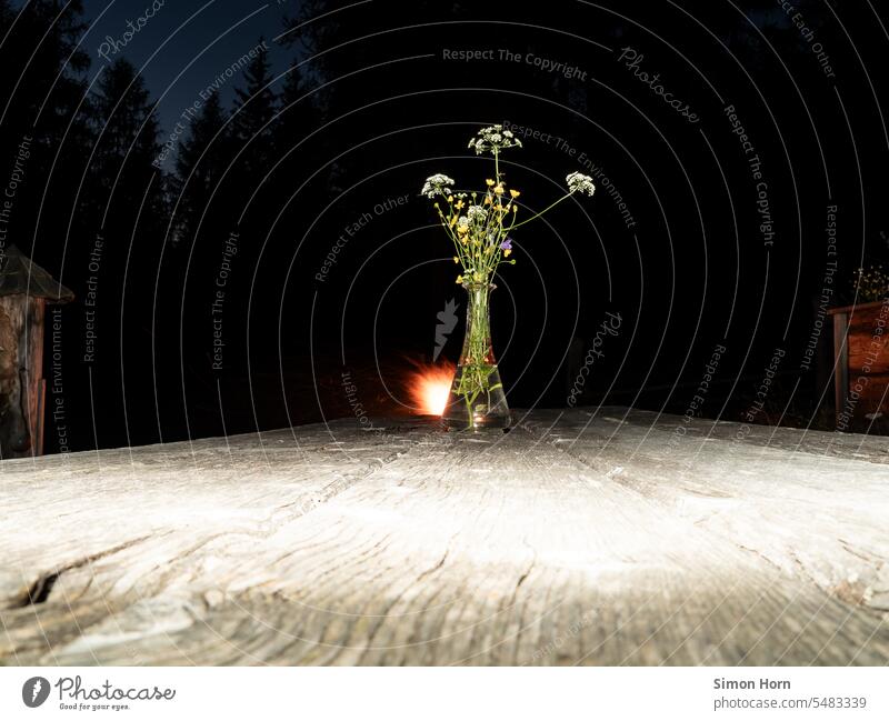 Wildblumenstrauß steht nachts auf einem Holztisch, im Hintergrund brennt ein Lagerfeuer Blumenstrauß Stimmung Nacht Dunkelheit Kontrast dunkel Feuer rustikal