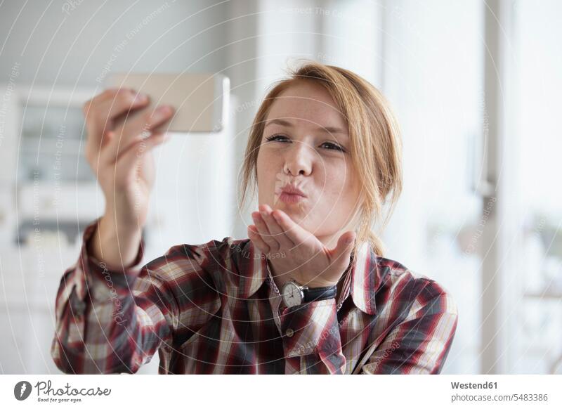 Junge Frau, die sich einen Kuss zuwirft, während sie ein Selfie mit ihrem Smartphone macht Hemd Hemden Haarsträhne Haarstraehne Strähne Selbstportrait Selfies