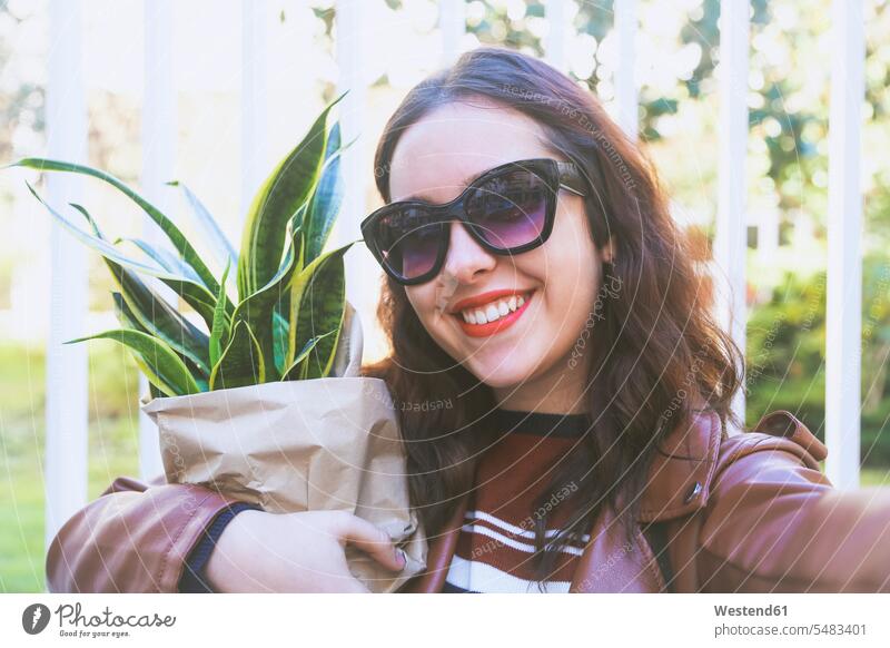 Porträt einer lächelnden jungen Frau mit Topfpflanze, die ein Selfie mit ihrem Smartphone macht Portrait Porträts Portraits weiblich Frauen Selfies Erwachsener