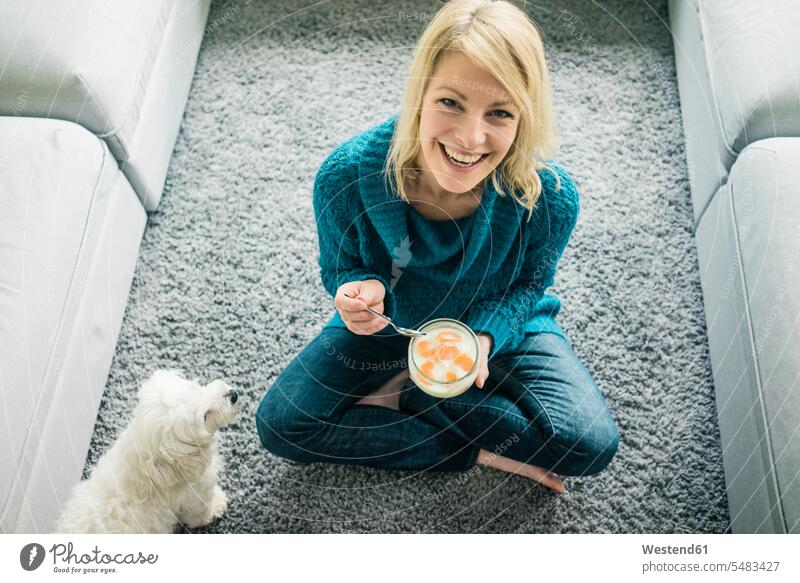 Porträt einer glücklichen Frau mit Hund beim Verzehr von Fruchtjoghurt im Wohnzimmer lächeln Hunde sitzen sitzend sitzt Joghurt essen essend weiblich Frauen