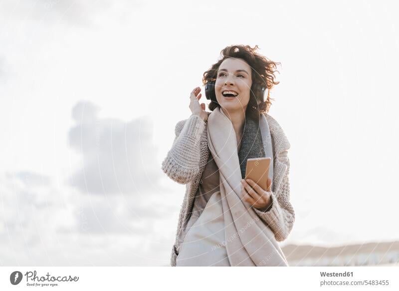 Glückliche Frau am Strand mit Handy und Kopfhörern Kopfhoerer Mobiltelefon Handies Handys Mobiltelefone Beach Straende Strände Beaches weiblich Frauen lachen