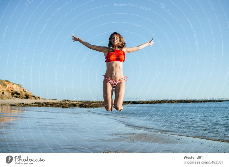 Lächelnde junge Frau springt am Strand in die Luft Beach Straende Strände Beaches Luftsprung Luftsprünge einen Luftsprung machen Luftspruenge weiblich Frauen