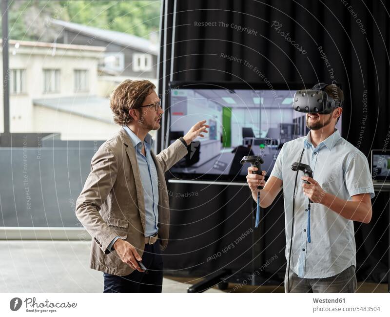 Mann spricht mit einem Kollegen, der eine VR-Brille vor dem Bildschirm trägt Brillen Arbeitskollegen Monitor Screen Monitore Bildschirme Screens