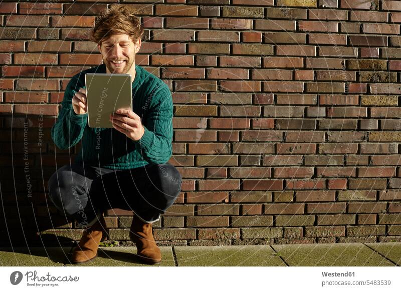Junger Mann kauert mit digitalem Tablett vor einer Ziegelmauer Europäer Kaukasier Europäisch kaukasisch Freude freuen Außenaufnahme draußen im Freien