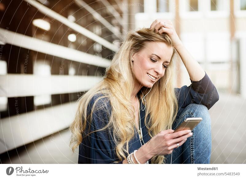 Porträt einer lächelnden blonden jungen Frau mit Kopfhörern, die auf ein Mobiltelefon schaut weiblich Frauen Portrait Porträts Portraits Erwachsener erwachsen