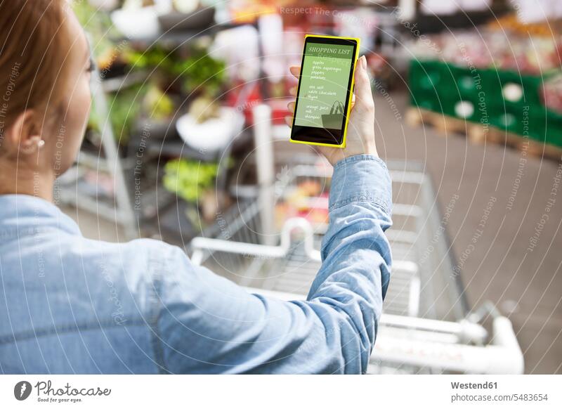 Frau hält ein Smartphone mit einer Einkaufsliste vor einem Supermarkt Liste Listen Supermaerkte Supermärkte einkaufen Einkaufen shoppen shopping Handy
