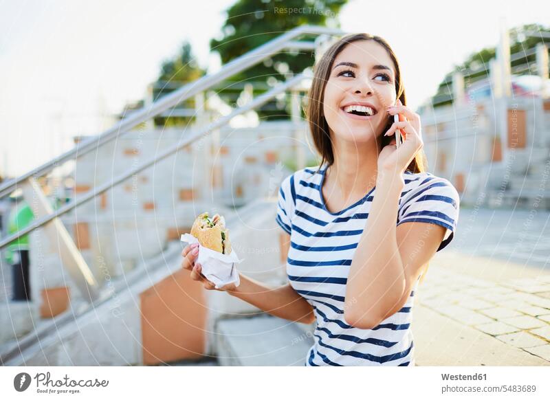 Junge Frau, die in der Stadt telefoniert und Bagel isst telefonieren anrufen Anruf telephonieren Handy Mobiltelefon Handies Handys Mobiltelefone lächeln