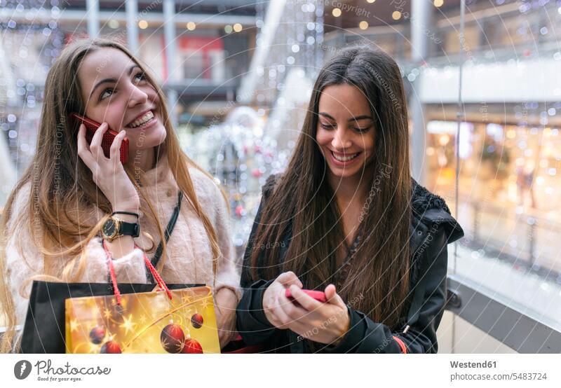 Zwei Freundinnen mit Einkaufstüten auf der Rolltreppe eines Einkaufszentrums mit ihren Smartphones Papiertüte Papiertüten offenes Lächeln lachen