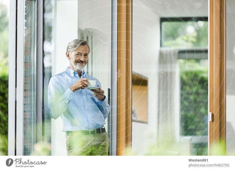 Lächelnder Mann mit Tasse Kaffee steht in seinem Haus und schaut durchs Fenster Männer männlich Erwachsener erwachsen Mensch Menschen Leute People Personen