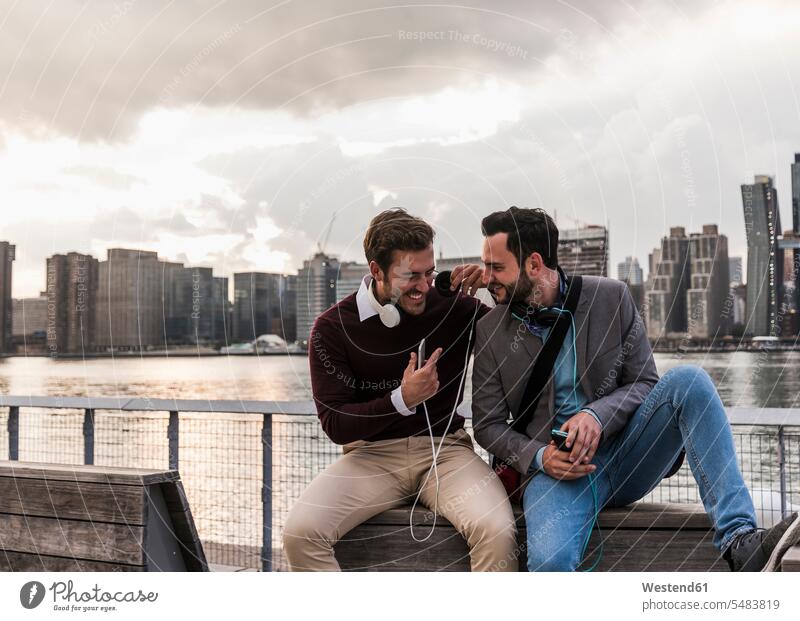 USA, New York City, zwei glückliche junge Männer mit Kopfhörern und Handy am East River sitzend lachen Freunde New York State Mann männlich positiv Emotion