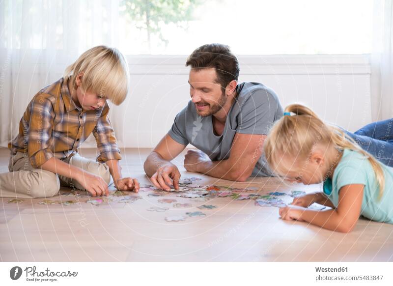 Vater mit zwei Kindern, die zu Hause auf dem Boden liegen und Puzzle spielen Böden Boeden liegend liegt Papas Väter Vati Vatis Papis Zuhause daheim Puzzlespiel