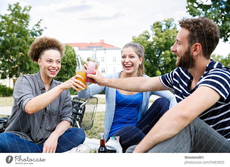 Drei glückliche Freunde klimpern mit Flaschen im Park anstoßen zuprosten anstossen lachen Glück glücklich sein glücklichsein Freundschaft Kameradschaft positiv