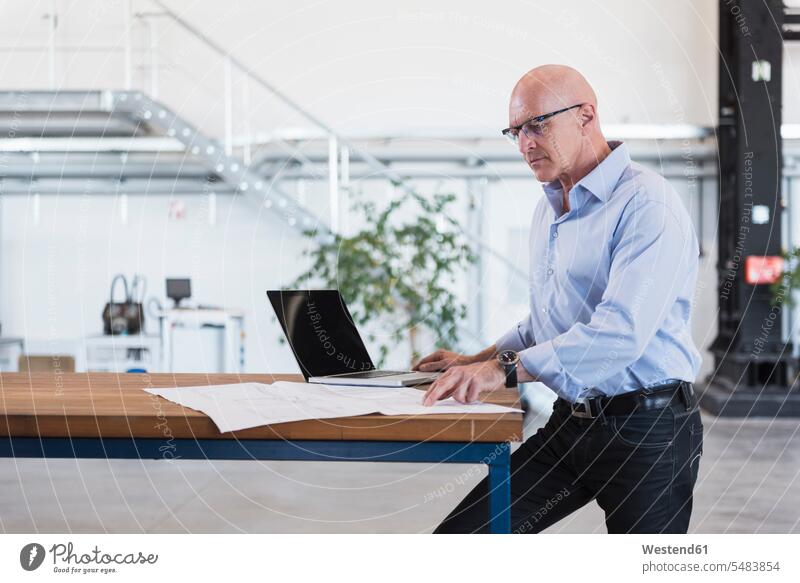 Mann mit Laptop schaut auf den Plan auf dem Tisch in der Fabrik Fabriken Geschäftsmann Businessmann Businessmänner Geschäftsmänner Notebook Laptops Notebooks