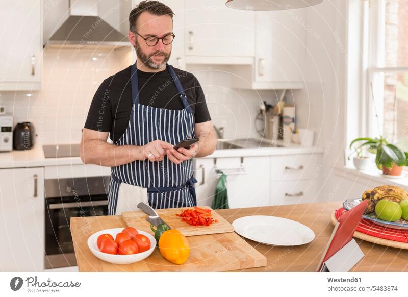 Porträt eines Mannes, der ein Smartphone benutzt, während er in der Küche Essen zubereitet simsen Paprika Gemuesepaprika Gemüsepaprika Paprikas Gemüsepaprikas