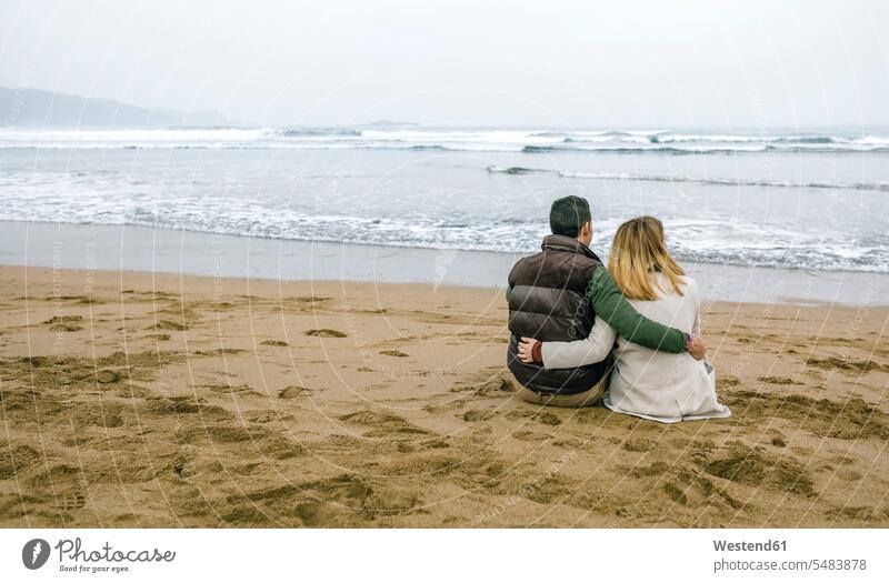 Verliebtes Paar sitzt im Winter am Strand Beach Straende Strände Beaches sitzen sitzend Pärchen Paare Partnerschaft Mensch Menschen Leute People Personen Meer