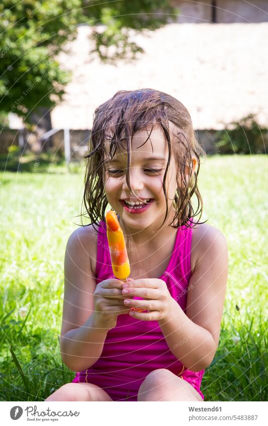 Porträt eines glücklichen kleinen Mädchens mit nassem Haar, das auf einer Wiese mit Eis am Stiel sitzt weiblich speisen essend Speiseeis Kind Kinder Kids Mensch