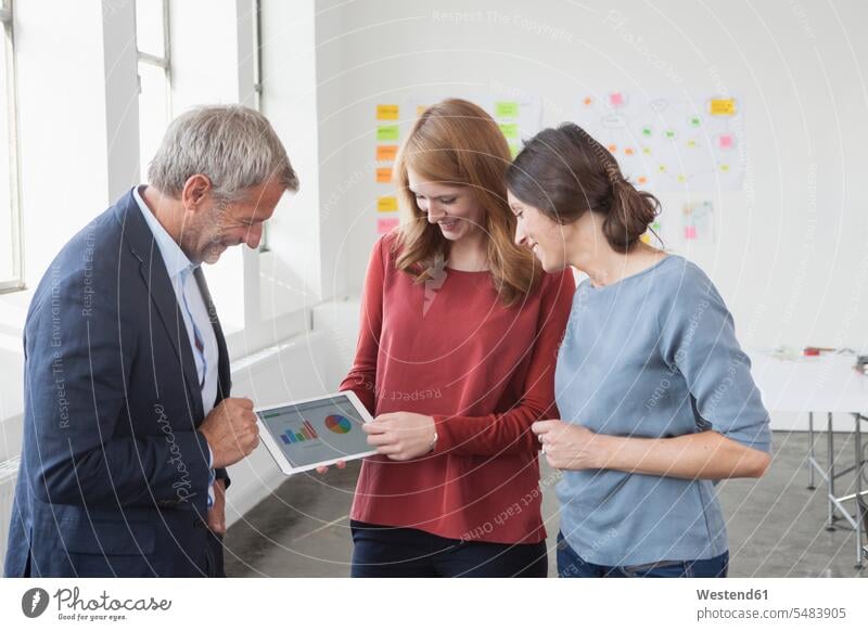 Lächelnder Praktiker und zwei Frauen im Büro schauen sich Diagramme auf digitalem Tablet an München Muenchen besprechen diskutieren Besprechung Meeting