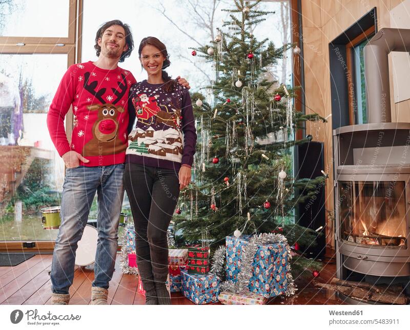 Paar, das vor dem Weihnachtsbaum steht und Weihnachtspullover trägt Europäer Kaukasier Europäisch kaukasisch Zusammengehörigkeit Verbundenheit Bindung