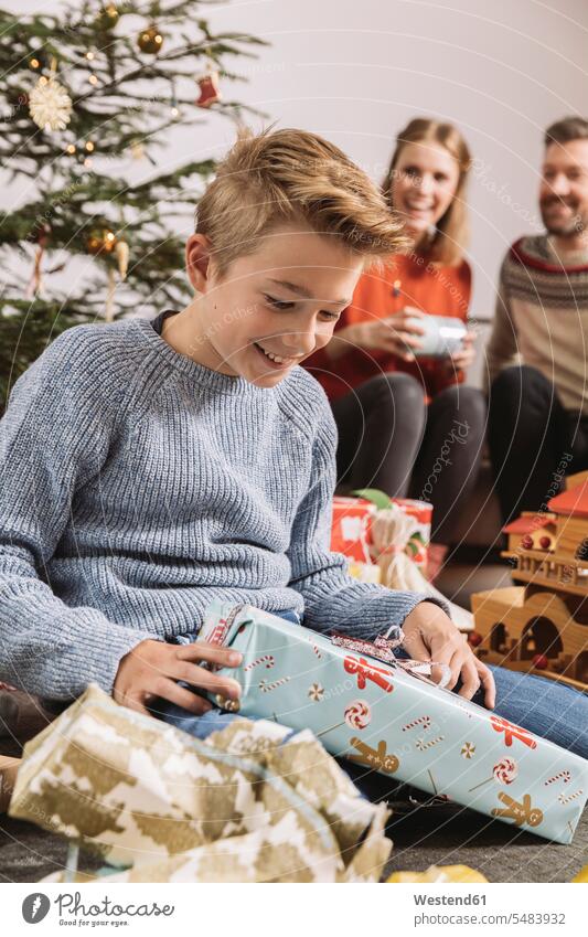Kleiner Junge packt ein Weihnachtsgeschenk aus, die Eltern sitzen im Hintergrund auf der Couch Europäer Kaukasier Europäisch kaukasisch Freude freuen Neugier