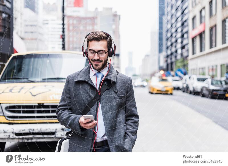 USA, New York City, Geschäftsmann mit Handy und Kopfhörern unterwegs Businessmann Businessmänner Geschäftsmänner lächeln Mann Männer männlich Kopfhoerer