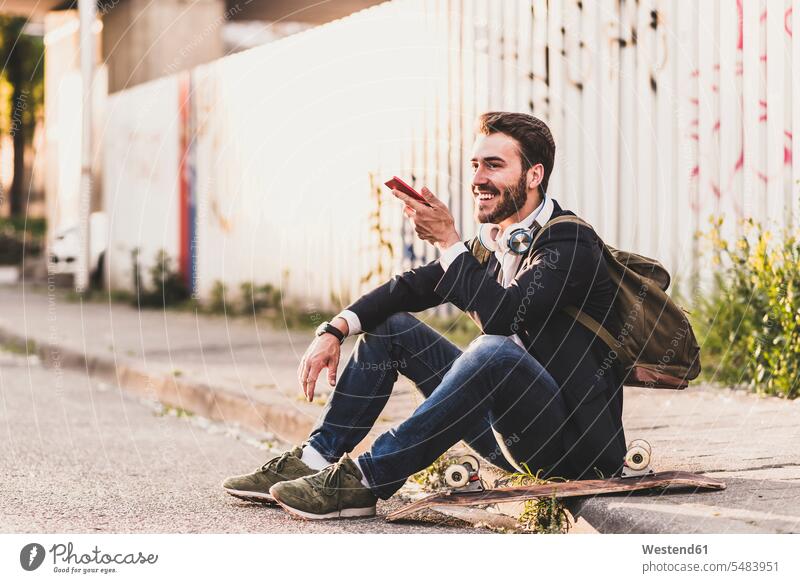 Lächelnder junger Mann sitzt auf dem Bürgersteig und benutzt sein Handy lächeln Mobiltelefon Handies Handys Mobiltelefone sitzen sitzend Männer männlich Telefon