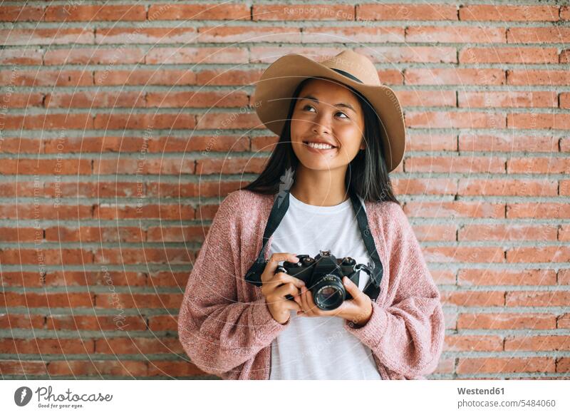Porträt einer lächelnden jungen Frau mit Kamera vor einer Ziegelmauer Kameras weiblich Frauen Fotoapparat Fotokamera Erwachsener erwachsen Mensch Menschen Leute