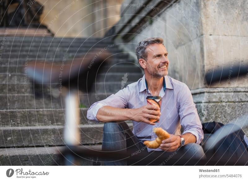 Lächelnder Mann auf der Treppe sitzend mit Croissant und Kaffee zum Mitnehmen in der Stadt lächeln Männer männlich Fahrrad Bikes Fahrräder Räder Rad sitzt