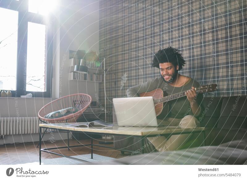 Mann sitzt im Wohnzimmer auf dem Sofa und spielt Gitarre vor dem Laptop Gitarren Männer männlich sitzen sitzend Notebook Laptops Notebooks Saiteninstrument