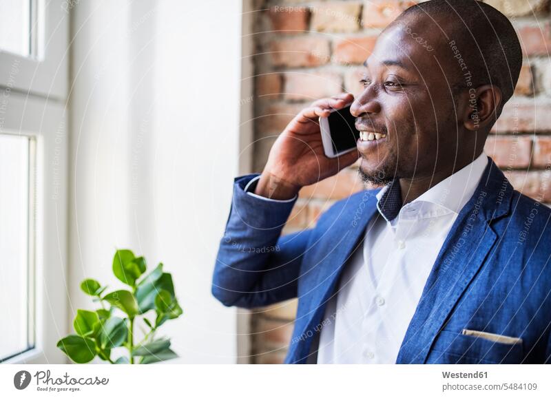 Lächelnder Geschäftsmann am Handy am Fenster Mobiltelefon Handies Handys Mobiltelefone Businessmann Businessmänner Geschäftsmänner telefonieren anrufen Anruf