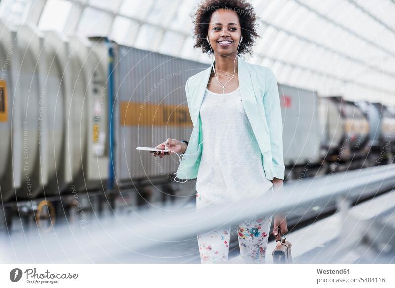 Porträt einer lächelnden jungen Frau mit Kopfhörern und Smartphone am Bahnsteig weiblich Frauen Erwachsener erwachsen Mensch Menschen Leute People Personen